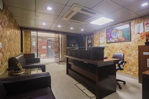Ein Empfangsbereich mit einer Rezeption, einer Sitzecke mit zwei Stühlen, einer Topfpflanze und einer Wand mit floralem Wandpapier sowie einem gerahmten Bild.