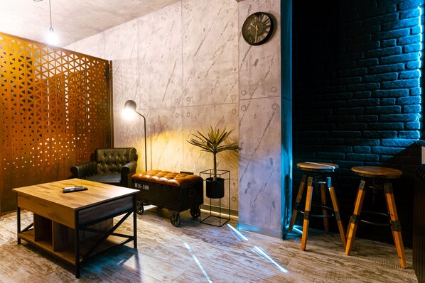 A modern lounge area with a leather armchair, a wooden table, a bar stool, a small table with a plant, and a wall clock.