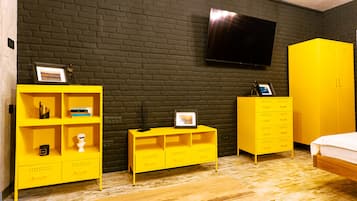 Huoneessa on tiiliseinä, keltainen vaatekaappi, jonka yläpuolella on seinälle asennettu litteänäyttöinen televisio, sekä valkoisella patjalla varustettu sänky.