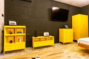A room with a brick wall, a flat-screen TV mounted above a yellow wardrobe, and a bed with a white mattress.