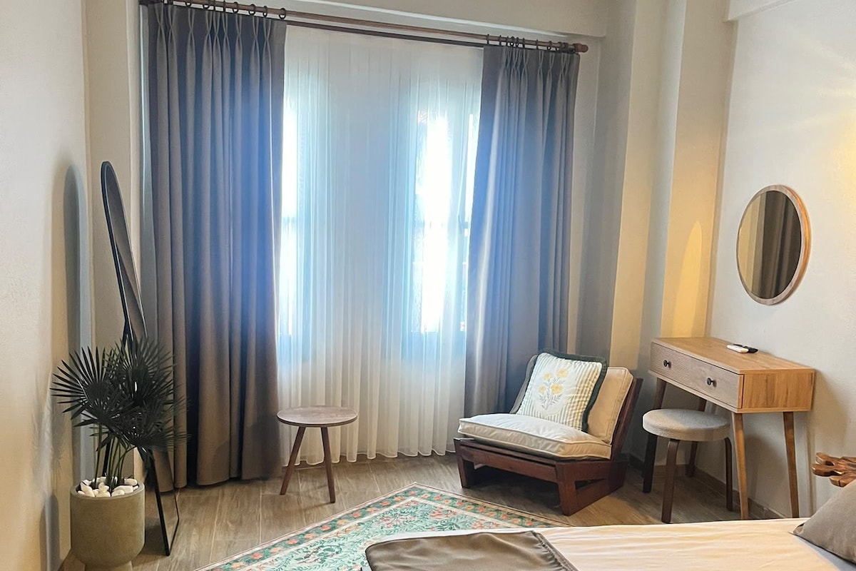 A bedroom with a bed, a desk, a chair, a potted plant, and a window with curtains.