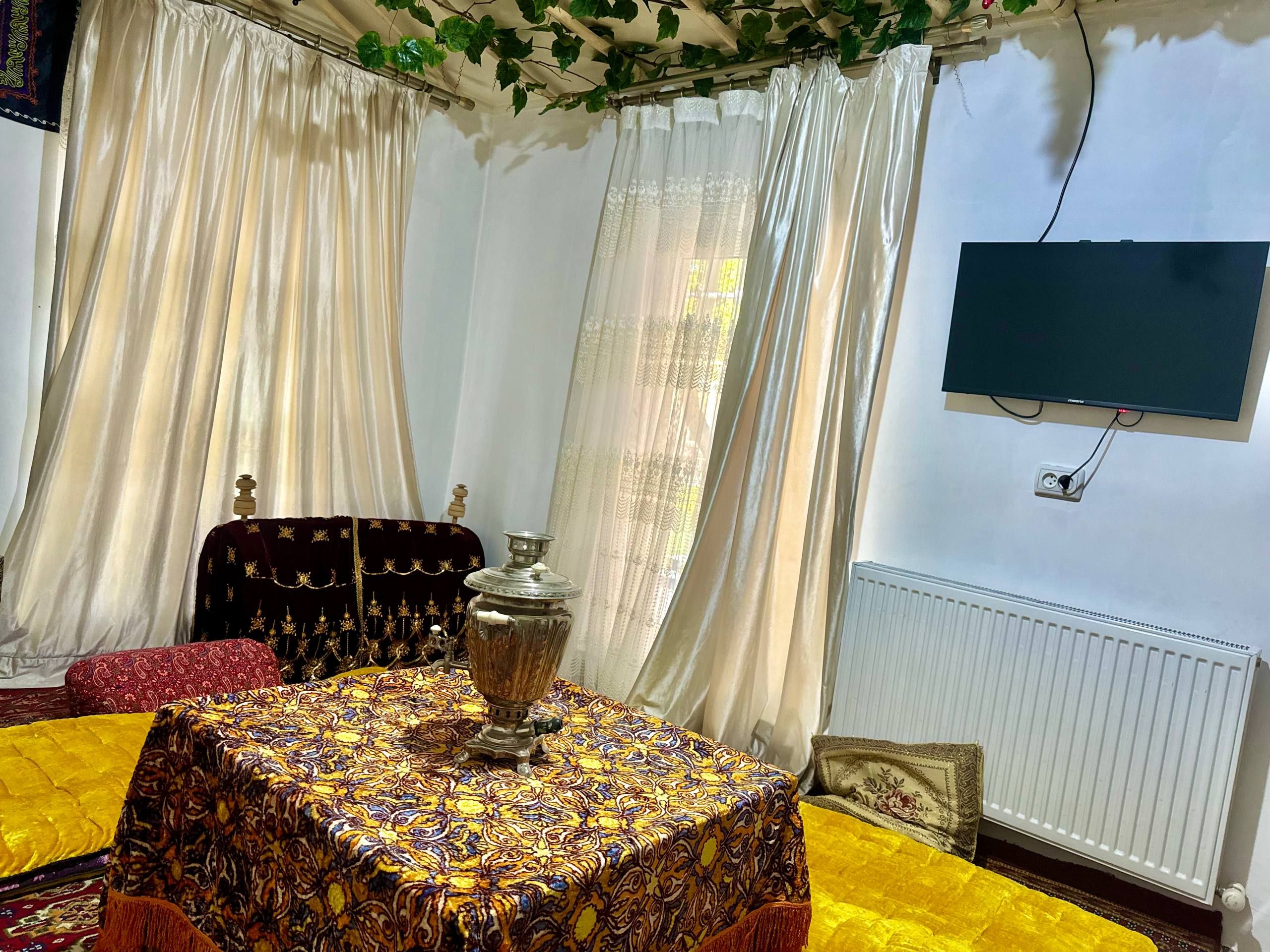 A room with a patterned tablecloth, a traditional coffee pot, a red sofa, and a television mounted on the wall.
