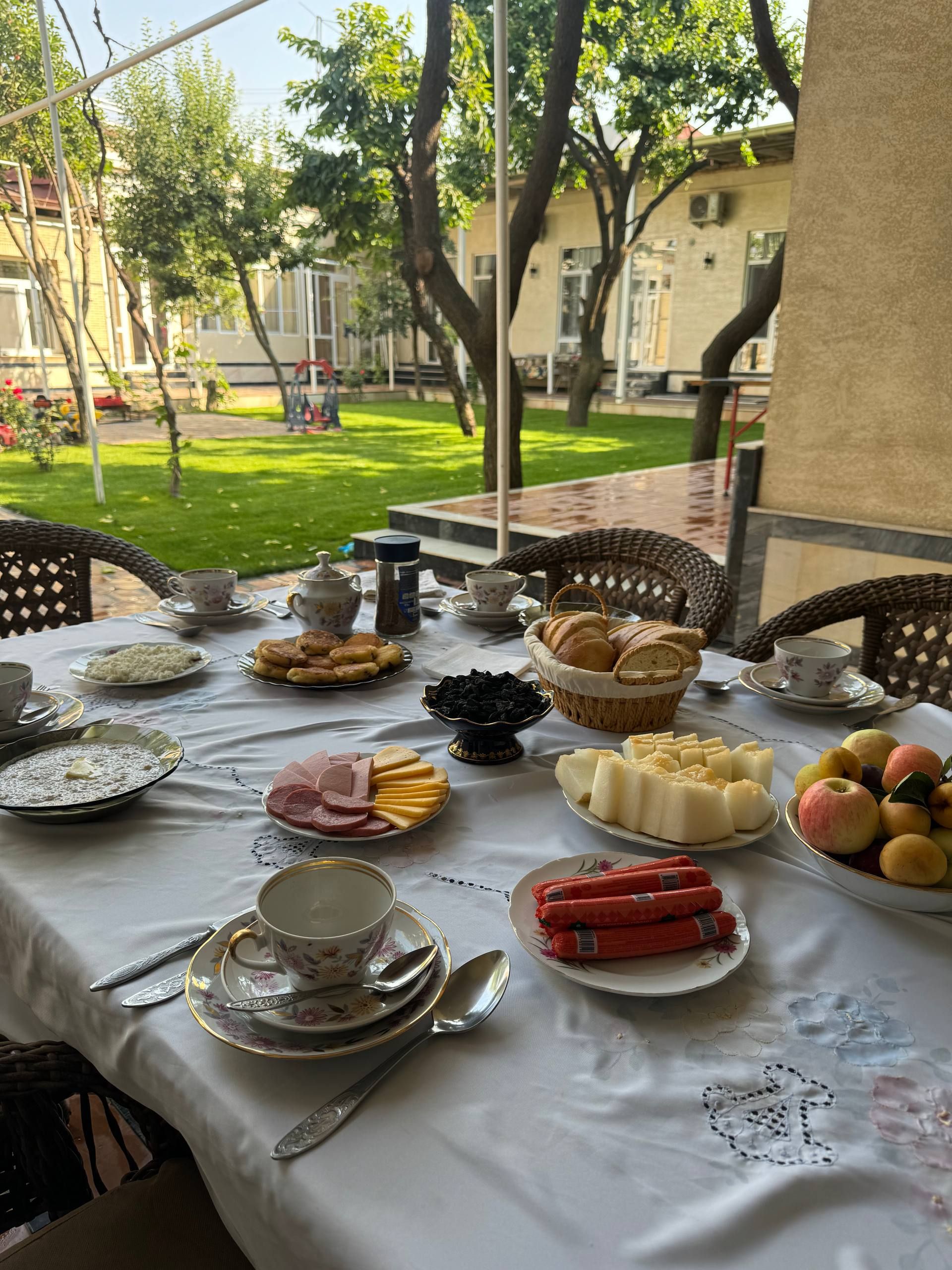 An outdoor dining area with a table set for a meal, surrounded by chairs, and a view of a garden and buildings in the background.