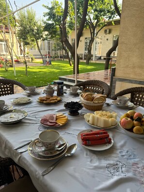 Uma área externa para refeições com uma mesa posta, rodeada por cadeiras, e vista para um jardim e edifícios ao fundo.