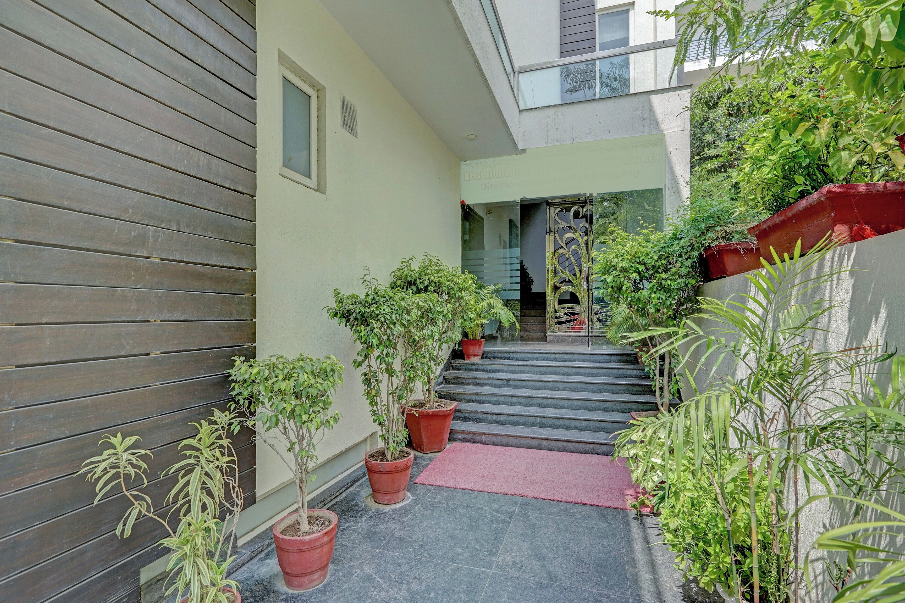A narrow pathway leading to a building entrance with potted plants and a red mat.
