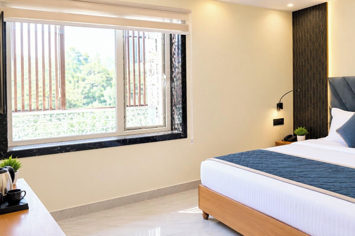 A hotel room with a large bed, a bedside table with a lamp, a window with blinds, and a view of greenery outside.