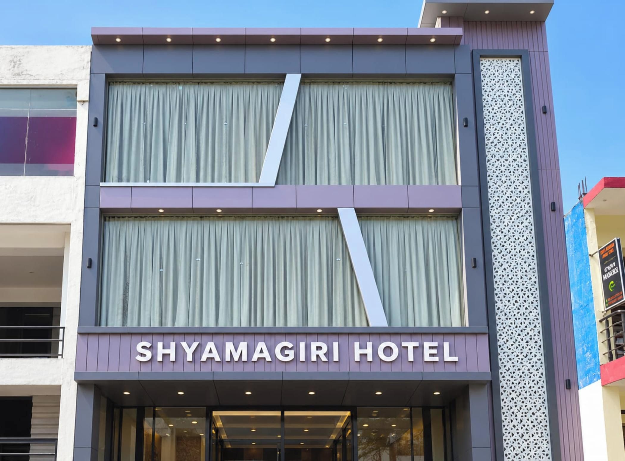 Exterior del Hotel Shyamagiri con fachada moderna, ventanas grandes y un llamativo letrero que dice 'Shyamagiri Hotel'.