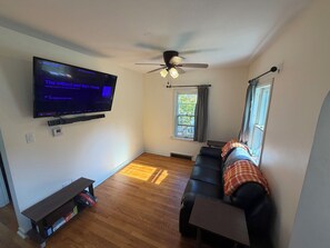 Olohuoneessa on seinälle asennettu taulutelevisio, kattotuuletin, ikkuna, jossa on verhot, musta nahkasohva ja puulattia.