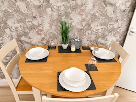 Mesa de madera redonda con cuatro platos blancos y posavasos negros, rodeada por dos sillas blancas.