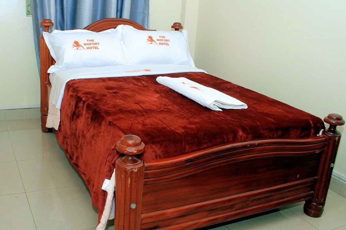 Un letto rifatto con una coperta rossa e cuscini bianchi, una testiera in legno e un asciugamano bianco piegato all'estremità del letto.