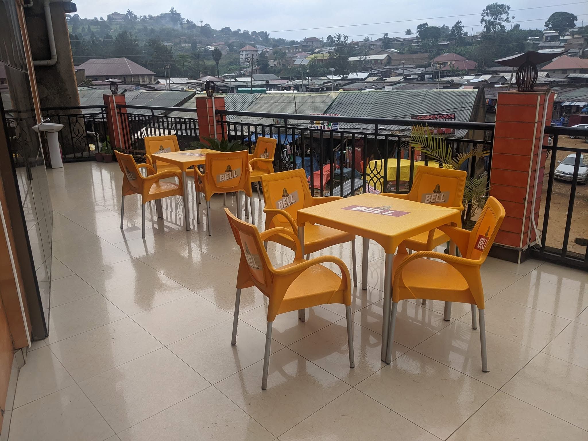 A terrace with yellow plastic chairs and tables, a tiled floor, and a view of a residential area with houses and a hill in the background.