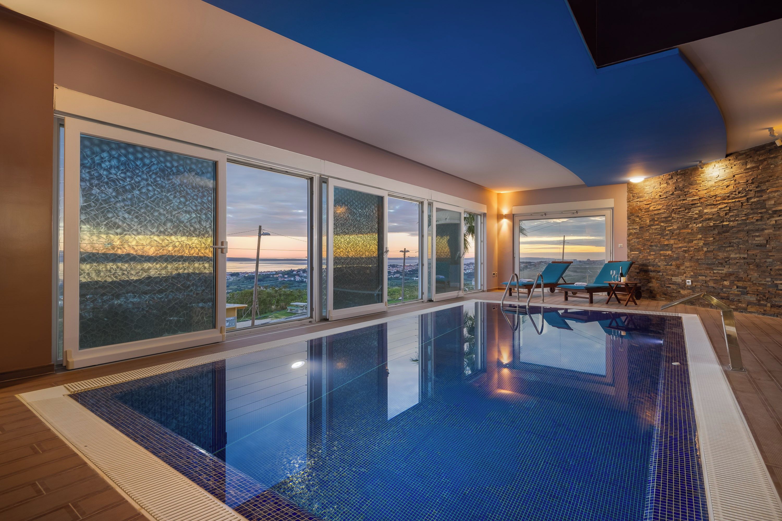 A modern indoor swimming pool with clear glass walls, a stone wall, and a sunset view.