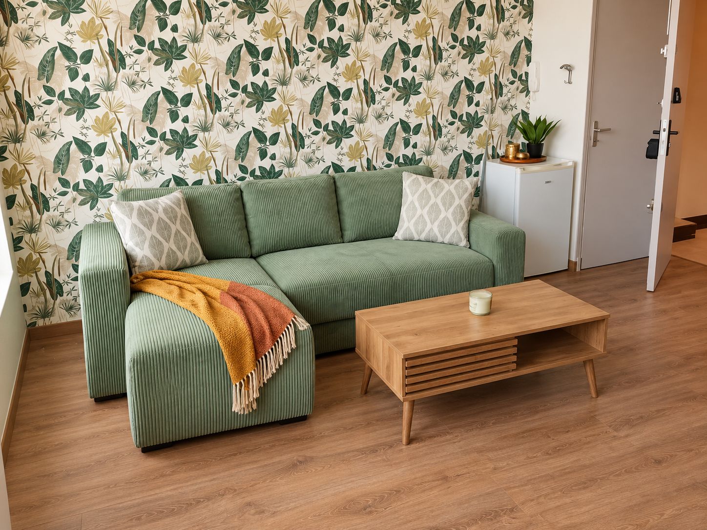 Ein grünes Sofa mit einer gemusterten Tagesdecke, ein Holztischchen und ein kleiner weißer Beistelltisch mit einer Pflanze.