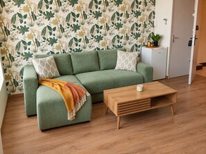 Un canapé vert agrémenté d’un plaid à motifs, une table basse en bois et une petite table d’appoint blanche sur laquelle est posée une plante.