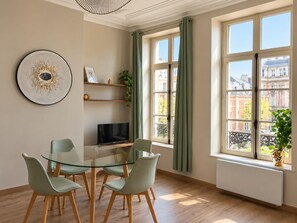 Ein modernes Wohnzimmer mit einem Glastisch, vier Stühlen, einem Flachbildfernseher und Blick auf Gebäude durch große Fenster.