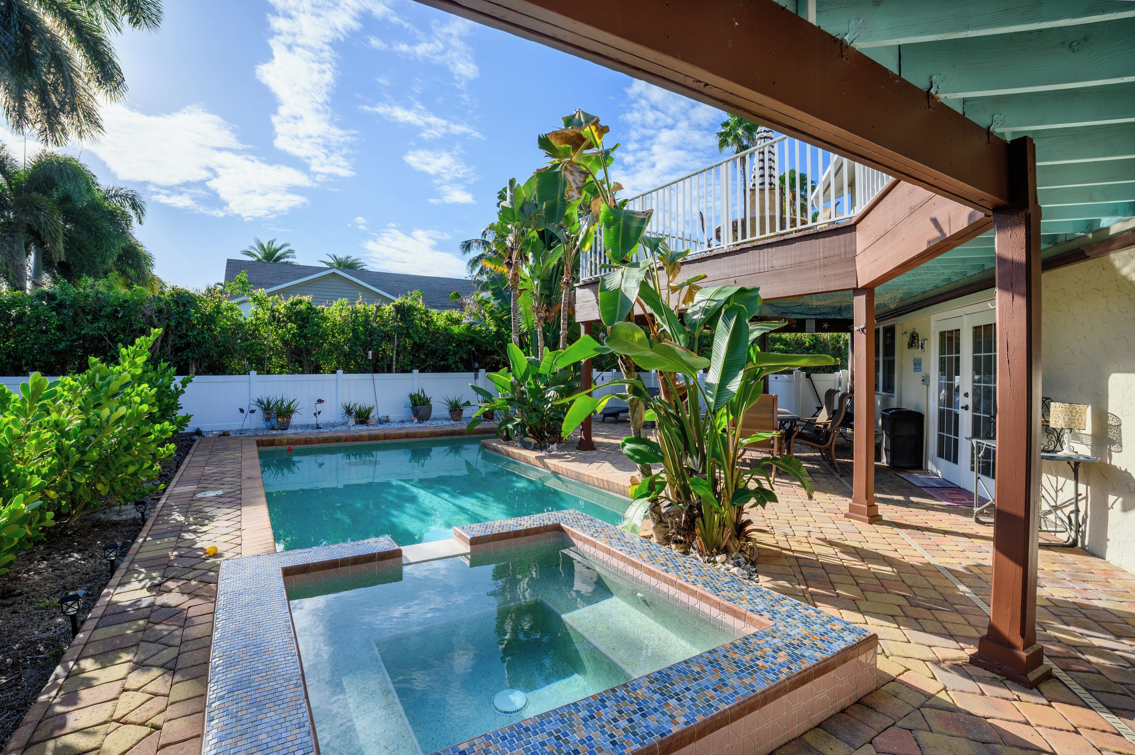 A swimming pool with a tiled deck, surrounded by lush greenery and a covered patio area.