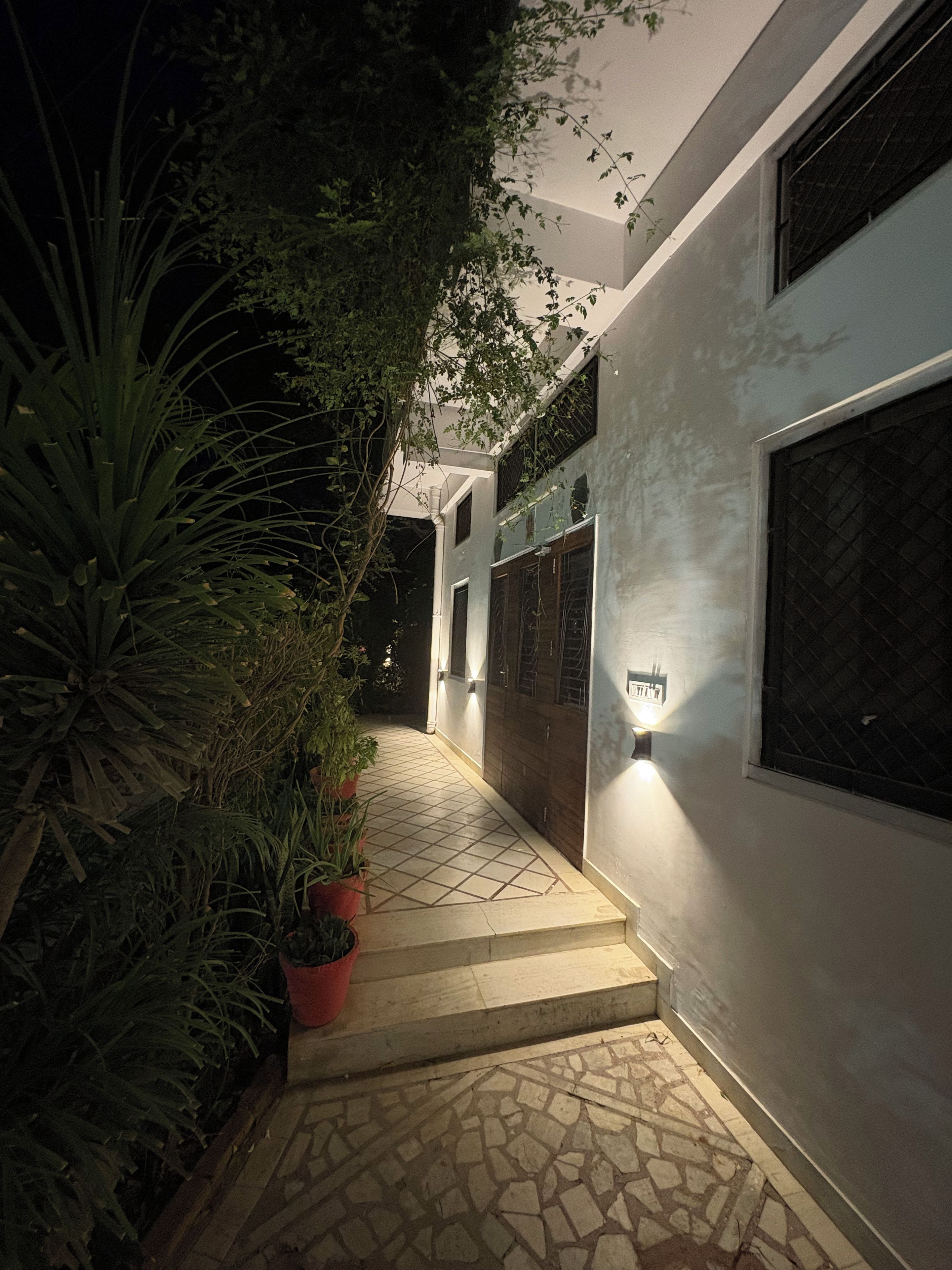 Vista nocturna del exterior de un edificio con un camino empedrado, plantas en macetas y un accesorio de luz empotrado iluminado.