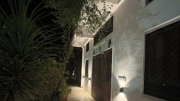 Vista nocturna del exterior de un edificio con un camino empedrado, plantas en macetas y un accesorio de luz empotrado iluminado.