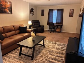 A living room with a brown leather sofa, a coffee table with a vase, a black leather chair, a wooden desk with a chair, and a television. - Serene 2-bedroom house with AC in charming Charleston location near all amenitie (Charleston)