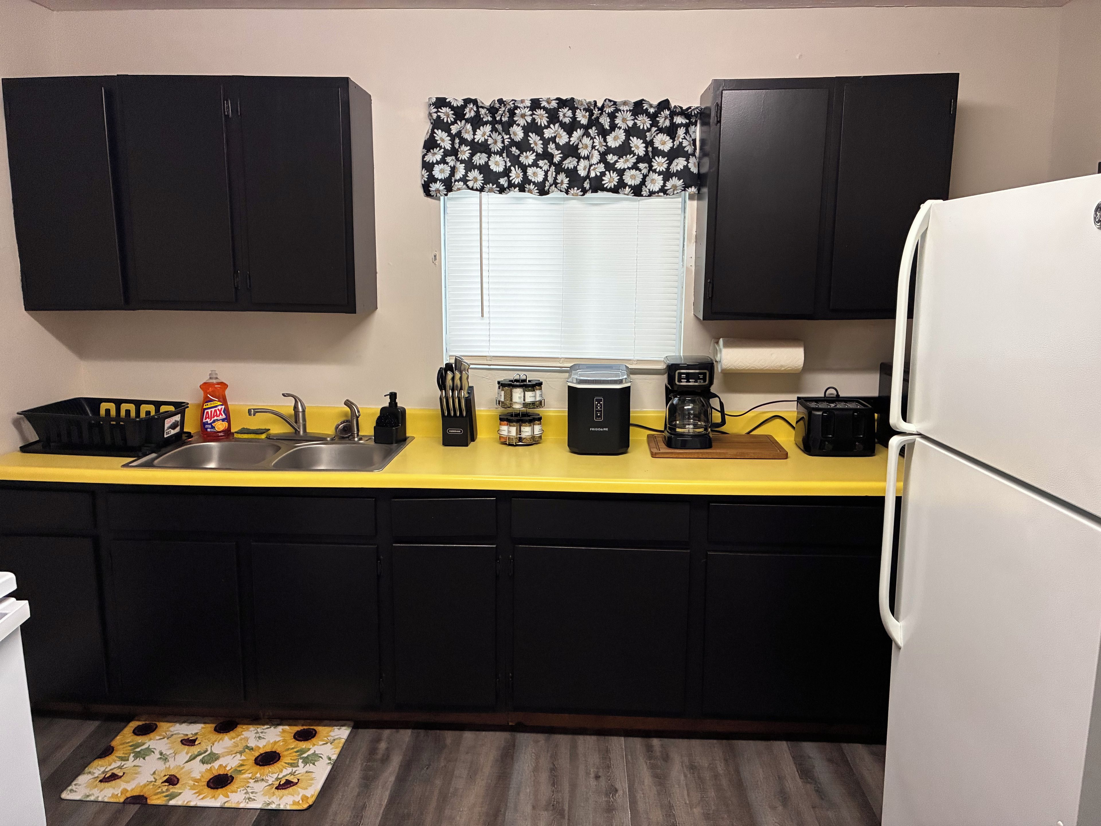 Cocina con armarios negros, encimera amarilla, cafetera y refrigerador.