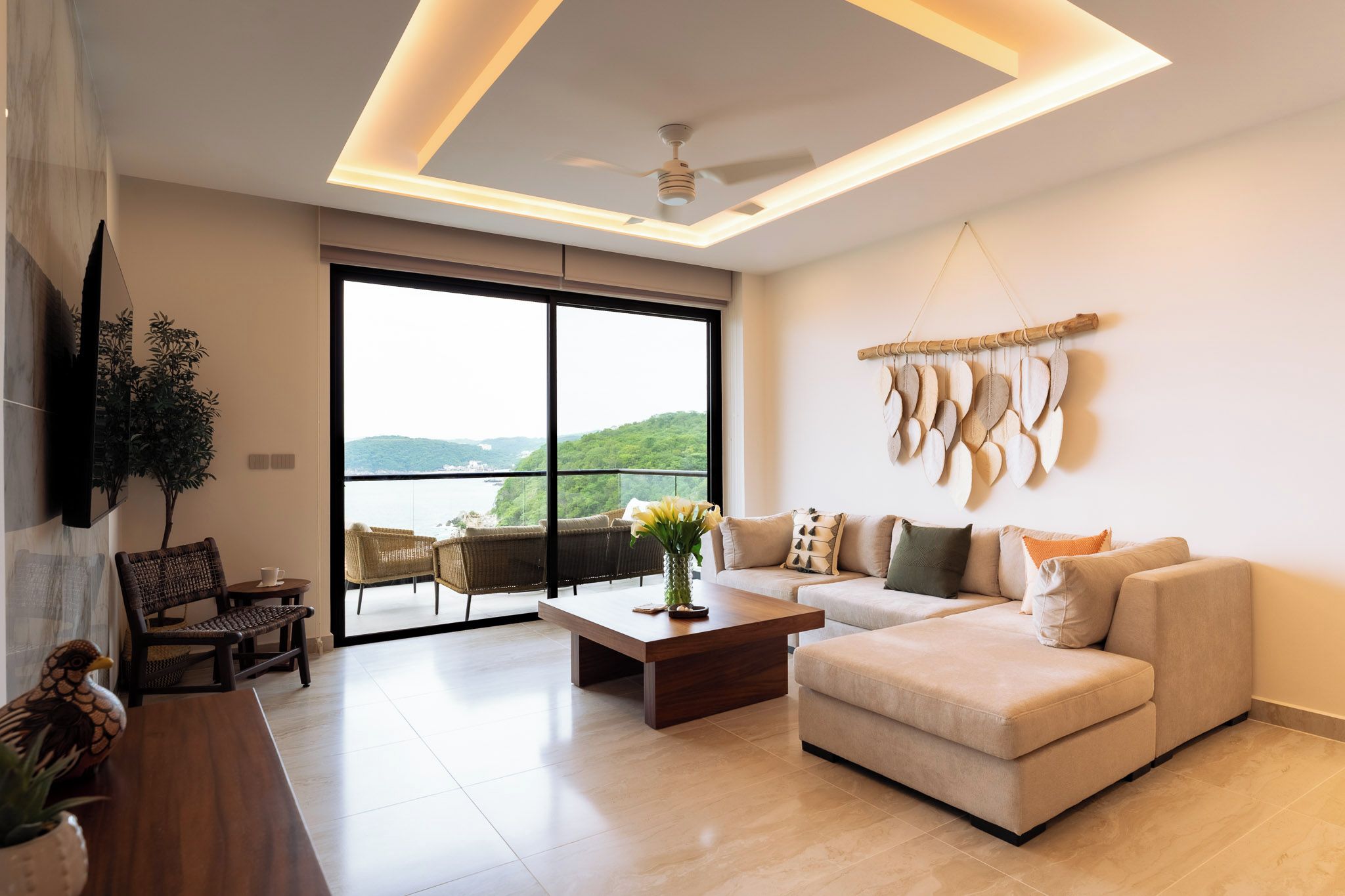A modern living room with a sofa, coffee table, and chairs. There is a large window with a view of greenery and hills.