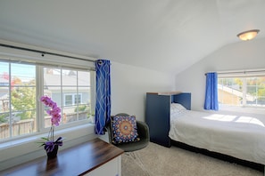 Quarto com cama, escrivaninha com um vaso de flores, cadeira e vista para o exterior.