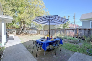 Um pátio externo com mesa posta para refeições, cercado por um muro de madeira e árvores.