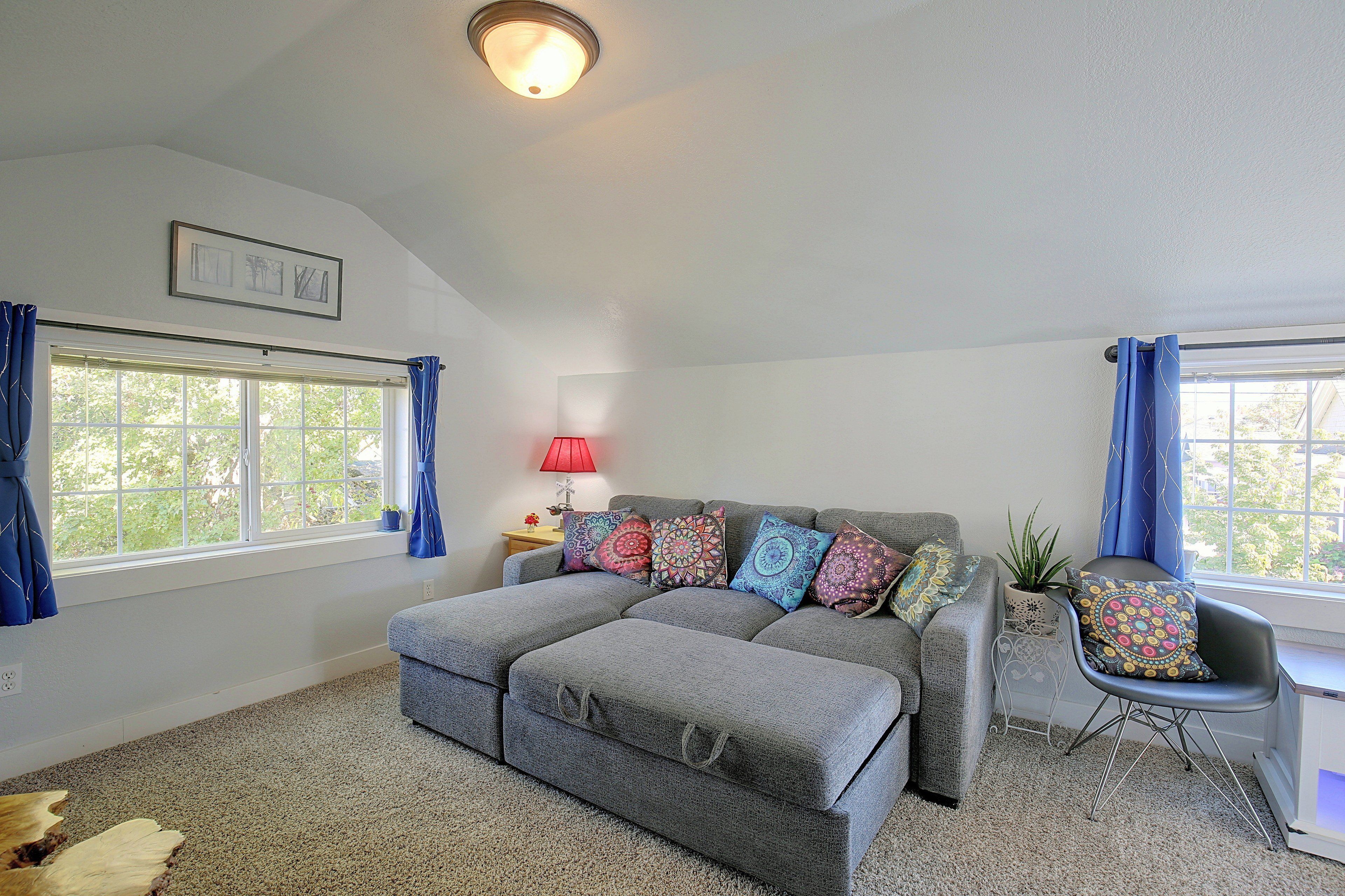 A living room with a grey sofa, a small side table, a red lamp, and a window with blue curtains.