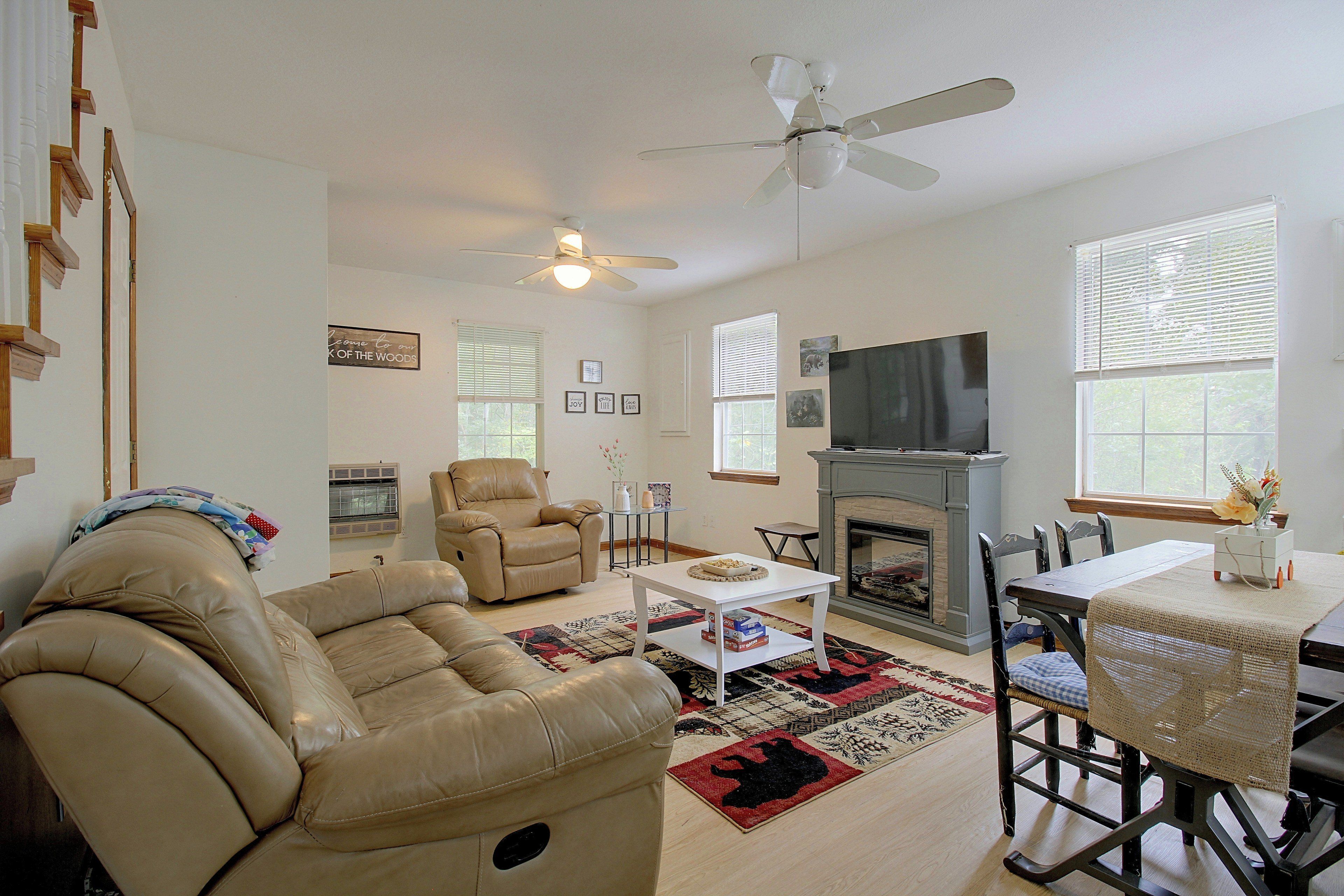 A living room with a fireplace, a television, a ceiling fan, and a large window with blinds.