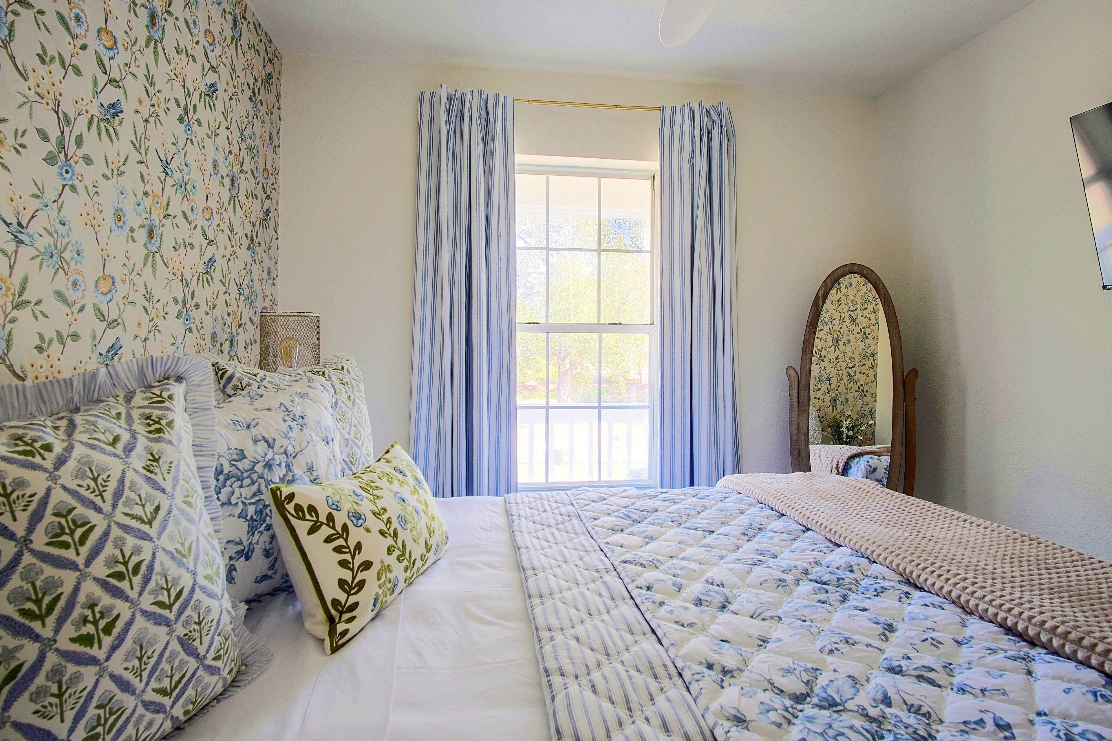 Una camera da letto con un'ampia finestra, un letto con biancheria a fiori e uno specchio sulla parete.