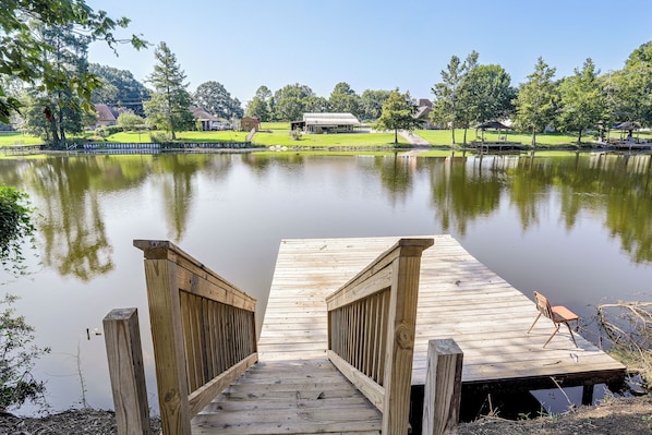 A wooden dock leading into a calm lake with a grassy park and buildings in the background.