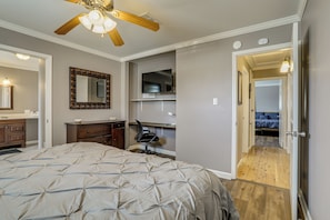 A bedroom with a bed, a desk, a chair, a ceiling fan, a mirror, and a door leading to another room.