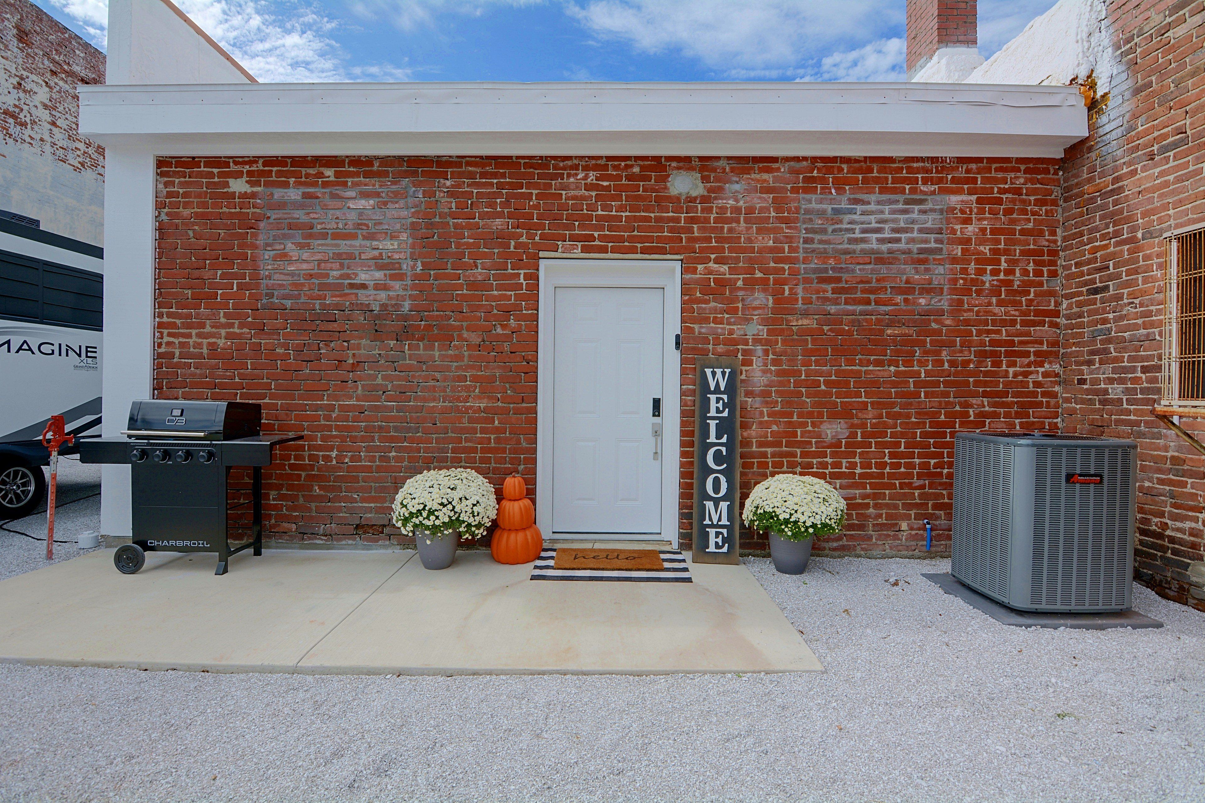 A brick building with a white door, a barbecue grill, two potted plants, a welcome sign, and an air conditioning unit.