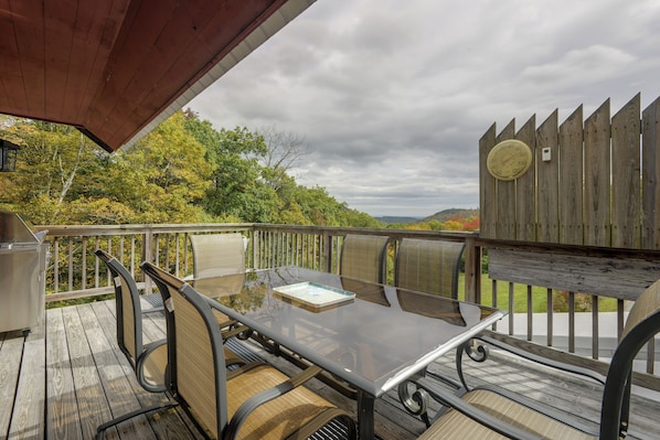 An outdoor deck with a glass-top table and chairs, a wooden fence, and a view of trees and hills.
