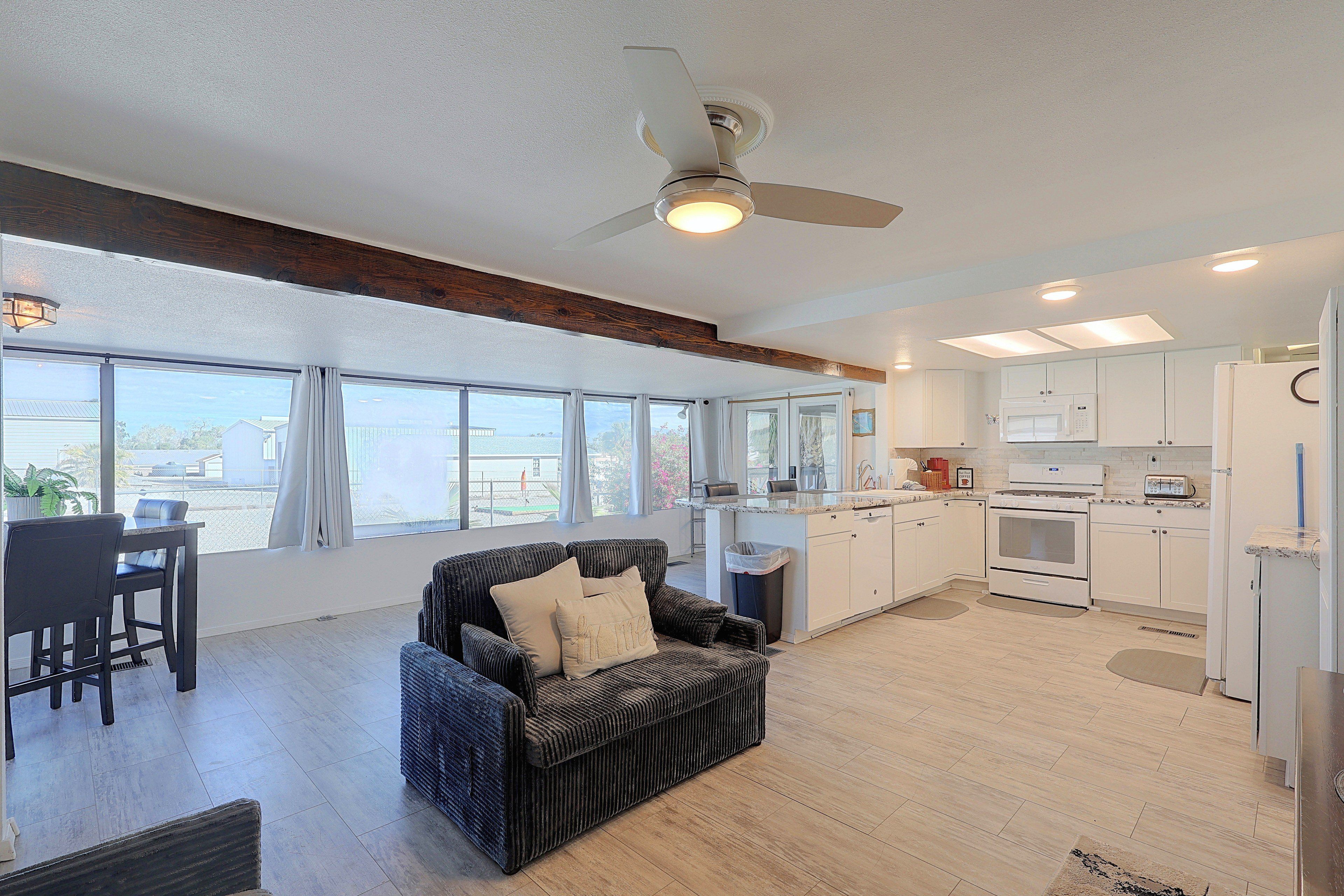 A spacious living area with a dark wicker sofa, a dining table with chairs, a kitchen with white appliances, and large windows offering a view of the outside.