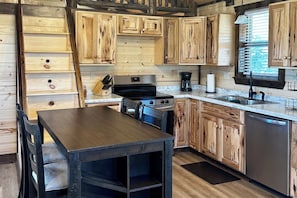 A kitchen with wooden cabinetry, a stove, a sink, and a dining area with a table and chairs. - Rimrock Ranch Retreat Near Fort Robinson Trails (Harrison)