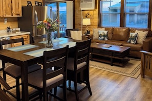 A modern kitchen with wooden cabinets, a stainless steel refrigerator, and a dining area with a wooden table and chairs. - Rimrock Ranch Retreat Near Fort Robinson Trails (Harrison)