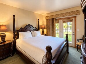 A hotel room with a large bed, two bedside tables with lamps, a wooden headboard, a window with curtains, and a view of the outdoors.