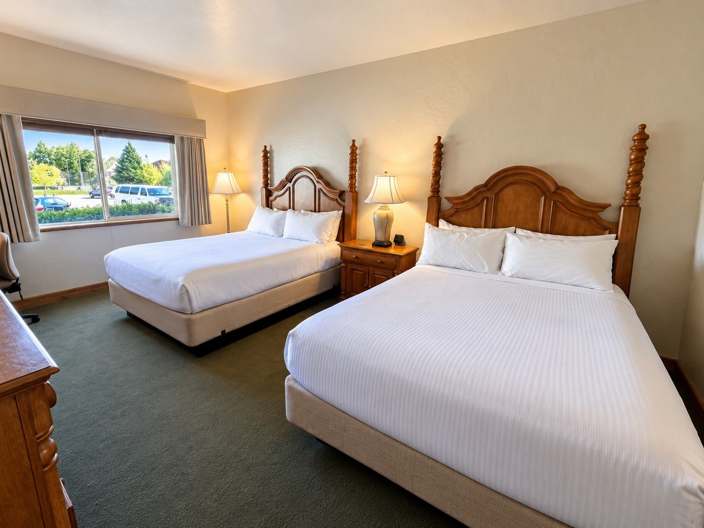 A hotel room with two beds, a desk, and a window with a view of parked cars and greenery.