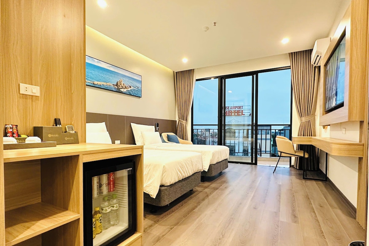 Habitación de hotel con cama, escritorio, minibar y balcón con vista al aeropuerto.