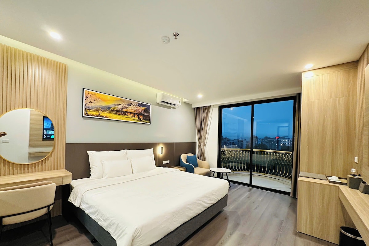 Una habitación de hotel moderna con una cama grande, un escritorio, una silla, un espejo redondo, un cuadro y un balcón con vista a la ciudad.