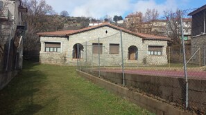 A stone building with a red-tiled roof, a fenced courtyard, and a hillside backdrop.