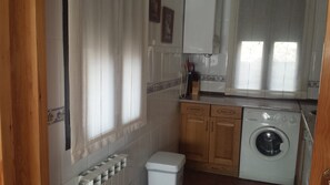 Eine kleine Küche mit Waschmaschine, Holzschränken, Heizkörper und einem Fenster mit Vorhängen.