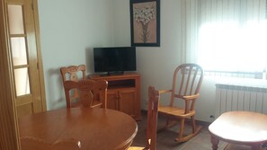 Ein Zimmer mit Esstisch, zwei Stühlen, einem Fernseher auf einem Schrank und einem Fenster mit Vorhängen.