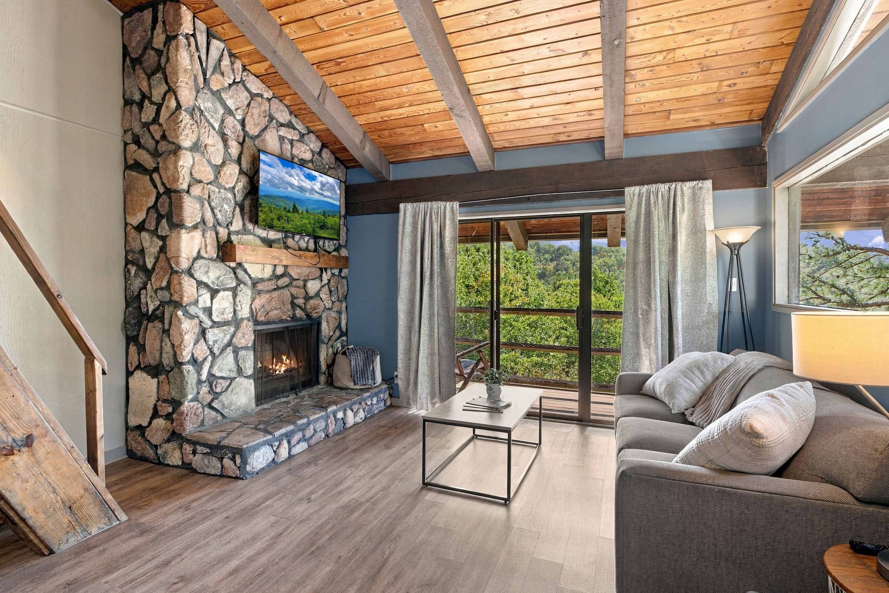 A cozy living room with a stone fireplace, a comfortable sofa, and a large window offering a view of greenery.