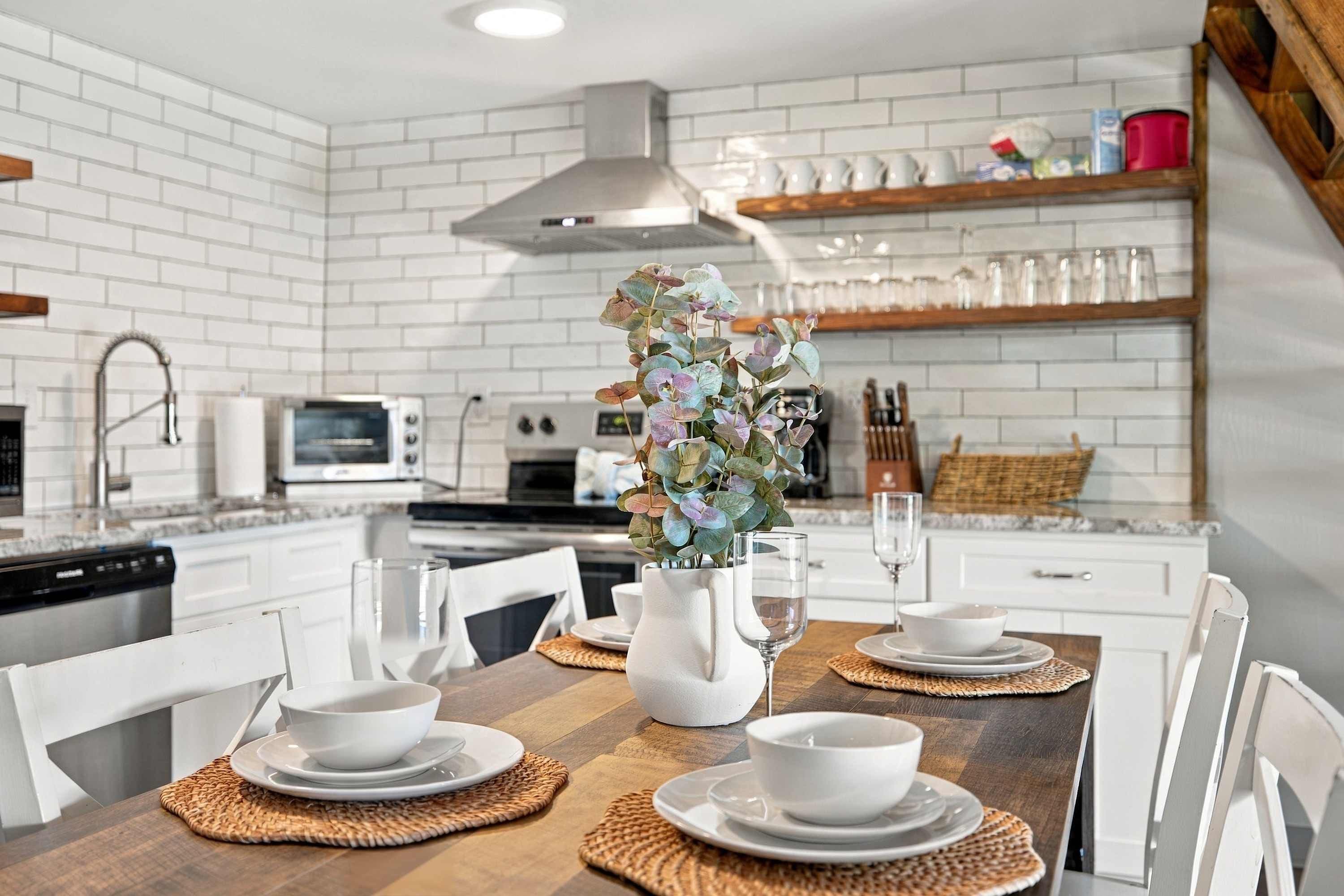 A modern kitchen with white brick walls, stainless steel appliances, and a dining area set with white plates, bowls, and a vase with flowers.