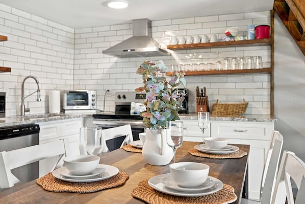 A modern kitchen with white brick walls, stainless steel appliances, and a dining area set with white plates, bowls, and a vase with flowers. - 2BR Top-floor Condo With Views Resort Amenities (Sevierville)