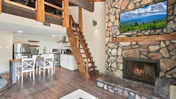 A spacious living area with a stone fireplace, a wooden staircase, and a kitchen in the background.