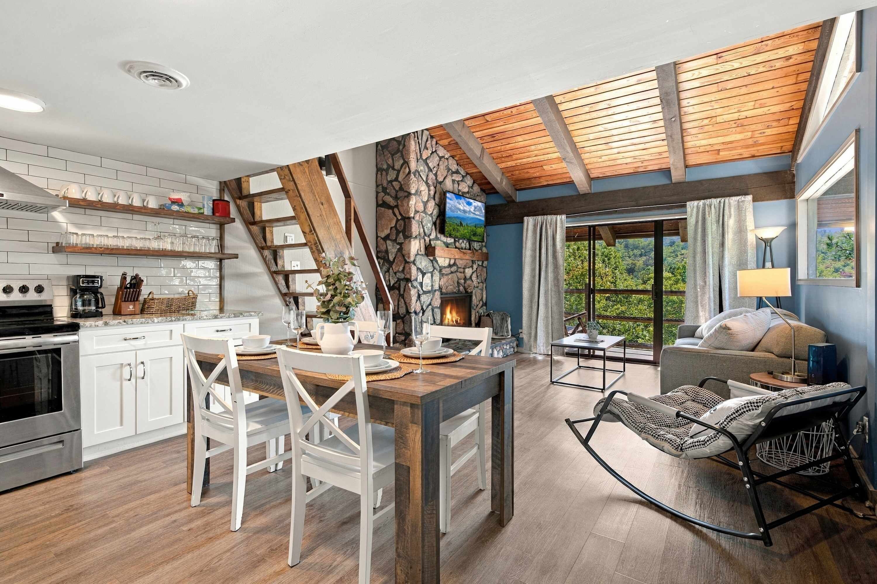 A modern kitchen with white cabinets, a dining area with a wooden table, and a stone fireplace.