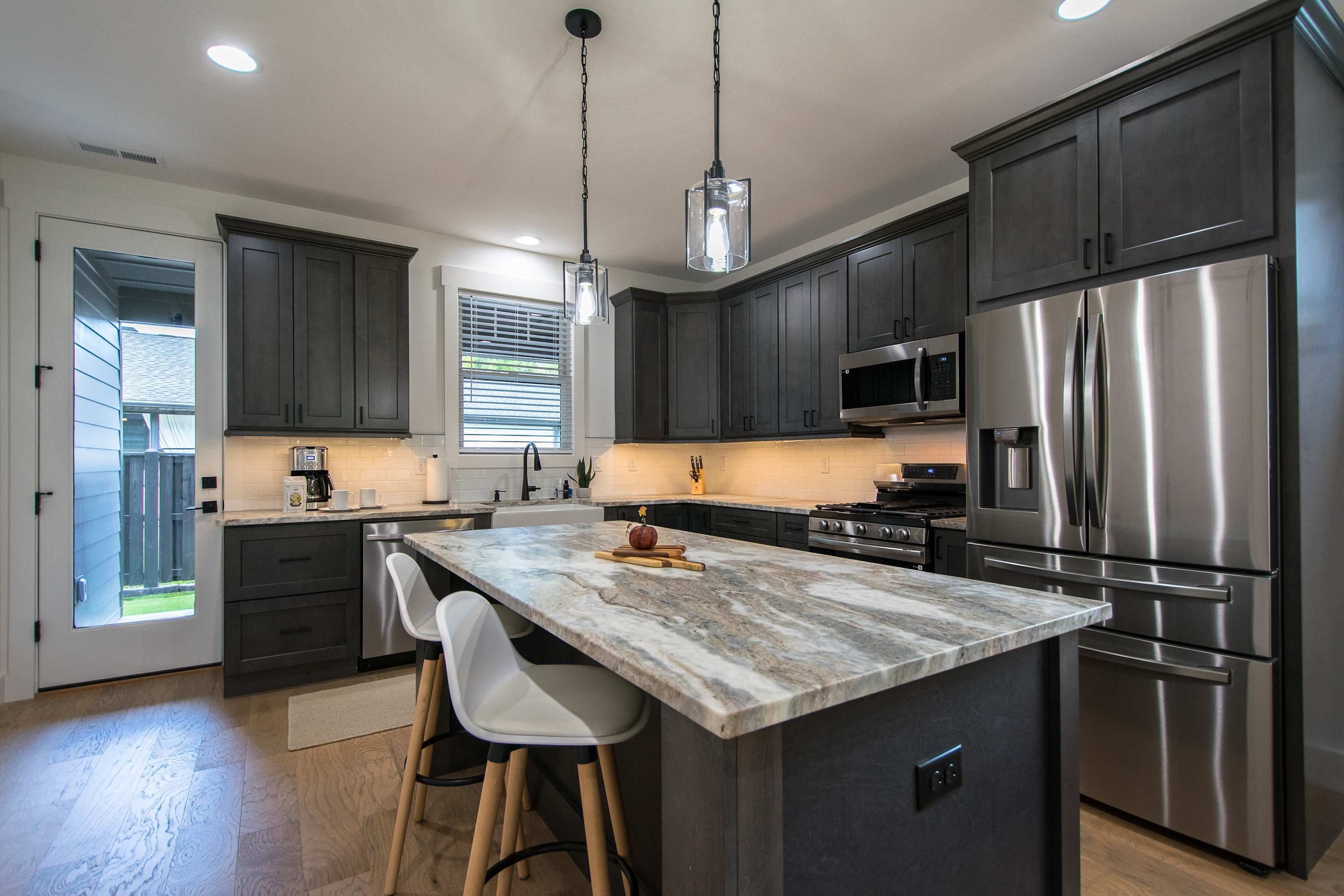 A modern kitchen with dark cabinets, stainless steel appliances, a marble island, and a pendant light.